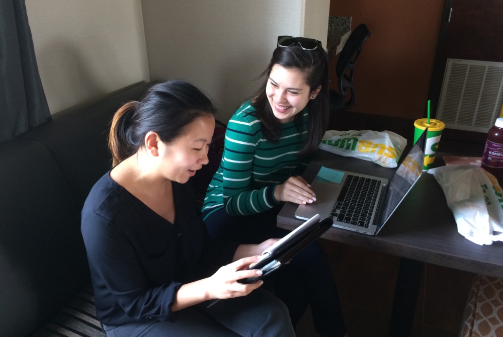 Two women working in front of a computer, one holding a notepad, are in conversation with one another