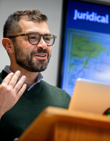 man speaking at a podium with a presentation on a screen behind him that reads 