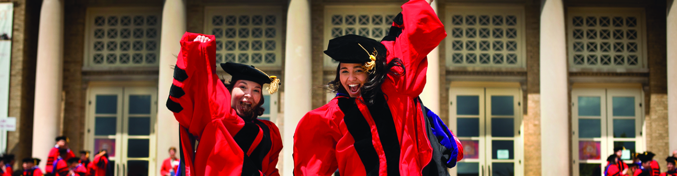 two young women in red graduation attire jump in front of a campus building