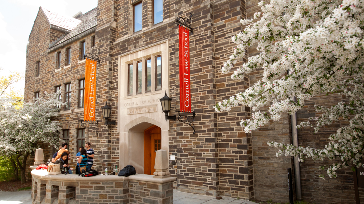 Students conversion near an entrance to Cornell Law School. Flowering trees are on both sides of the image, and vertical banners displaying "Cornell Law School" flank the building entrance.