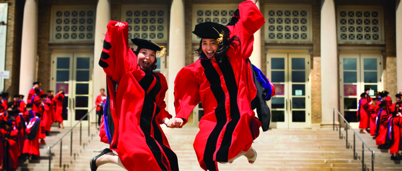 two young women in red graduation attire jump in front of a campus building