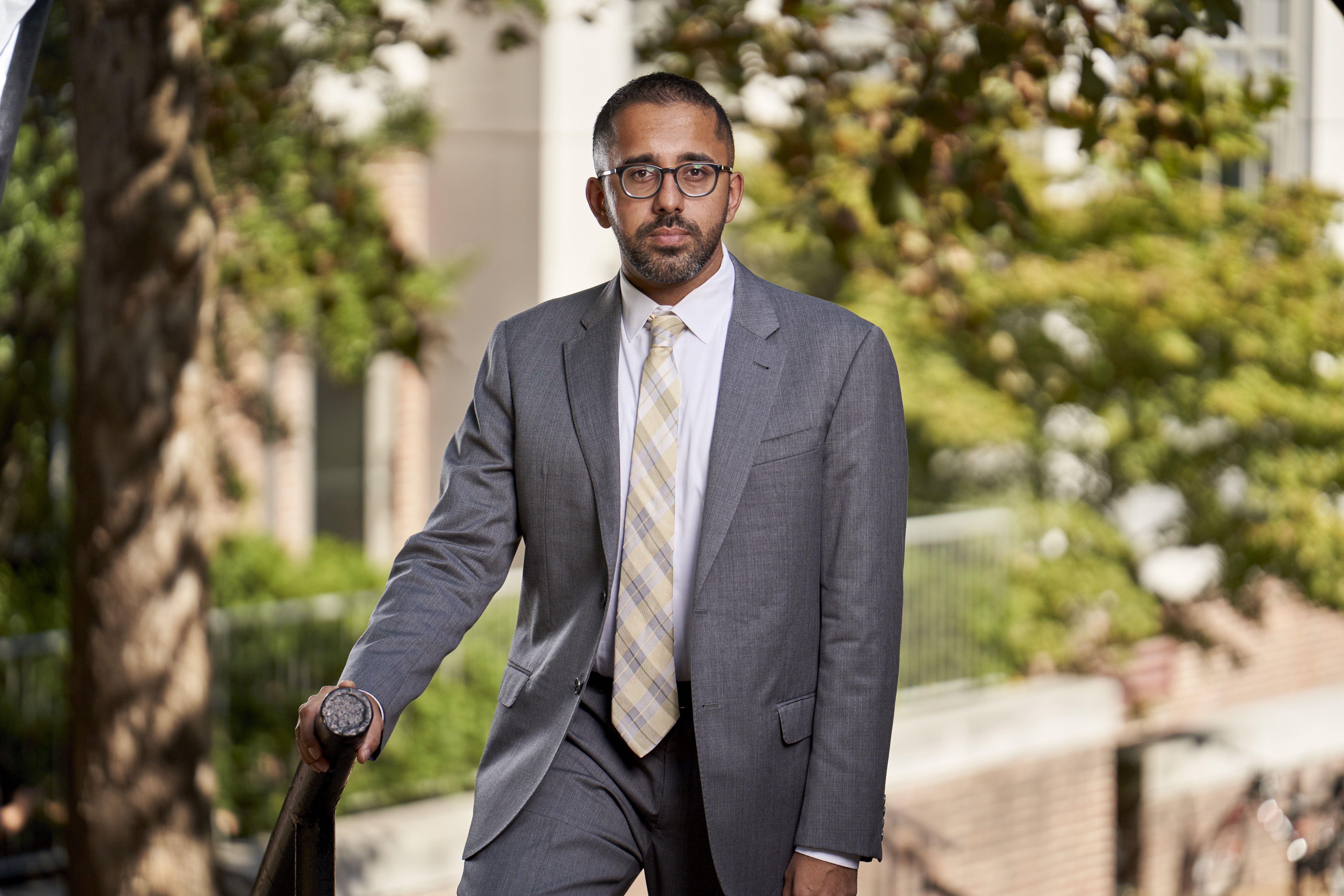 Photo of Gautam Hans standing outside in a grey suit, resting his hand on a railing. Trees are blurred in the background