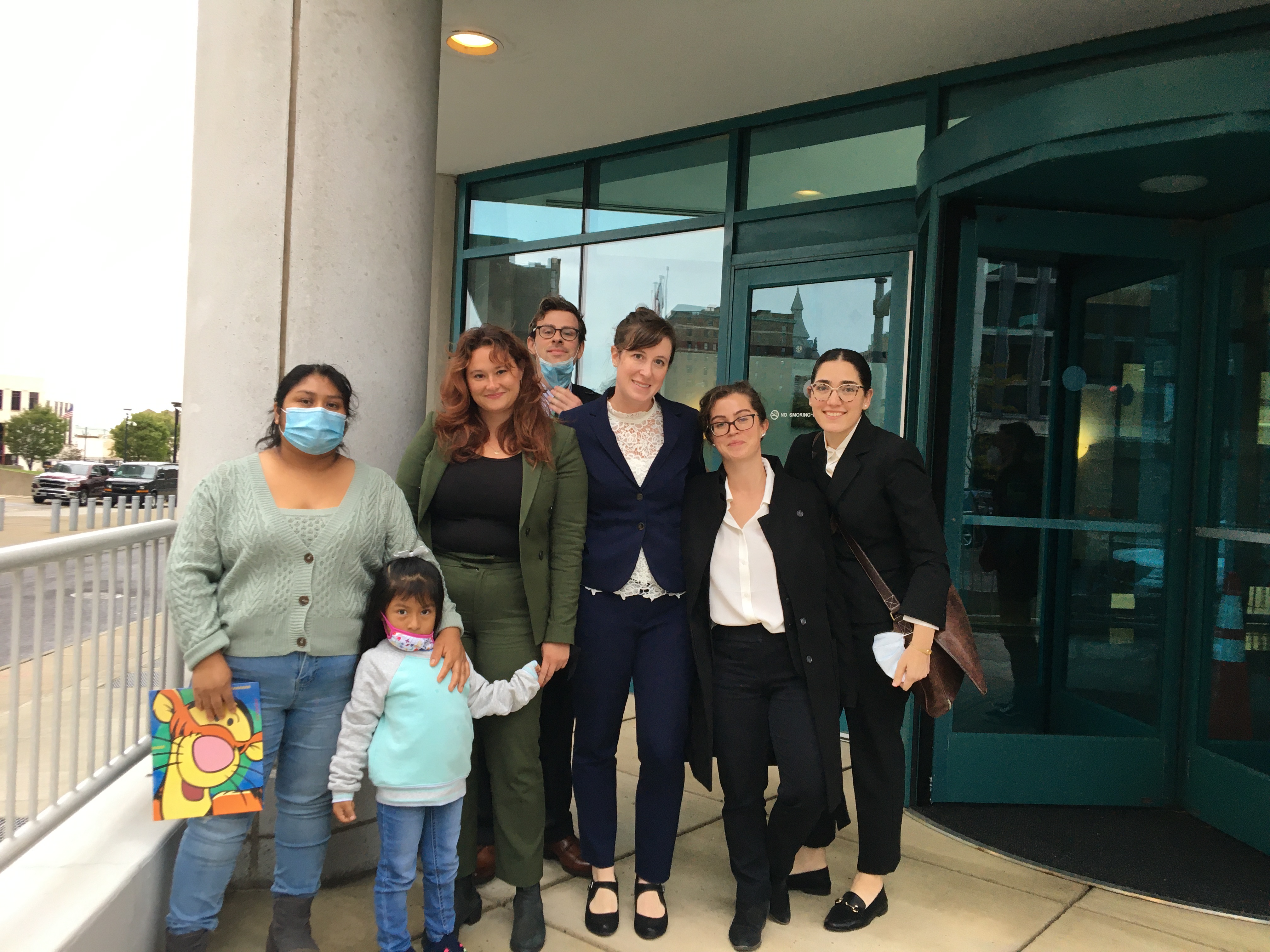 Photo of several members of the legal team standing with their client and their two small children, outside in front of a building's entrance.