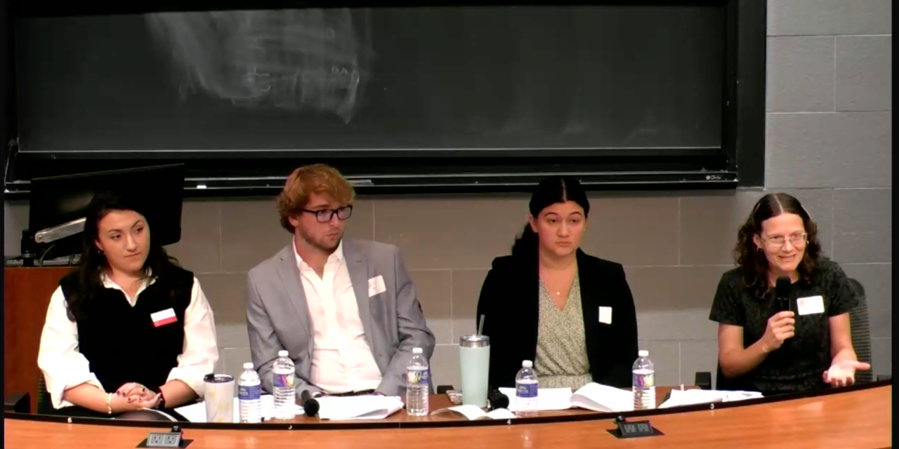 Photo of students sitting for a panel behind a long wooden table with table top mics