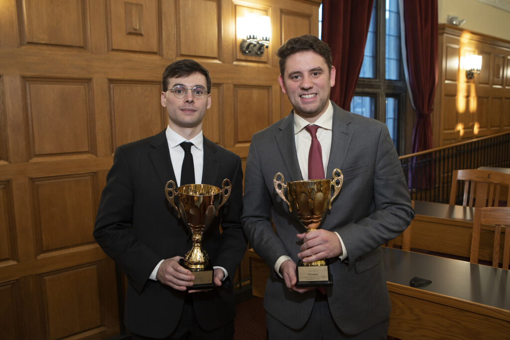 Two gentlemen staring at the camera wearing suits and holding gold trophies for winning the moot court competition.