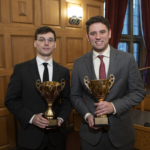 Two gentlemen staring at the camera wearing suits and holding gold trophies for winning the moot court competition.
