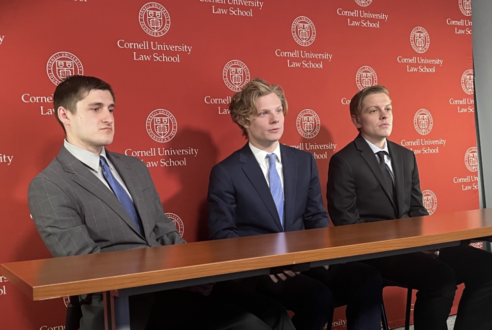 three men in suits seated in front of a Cornell banner