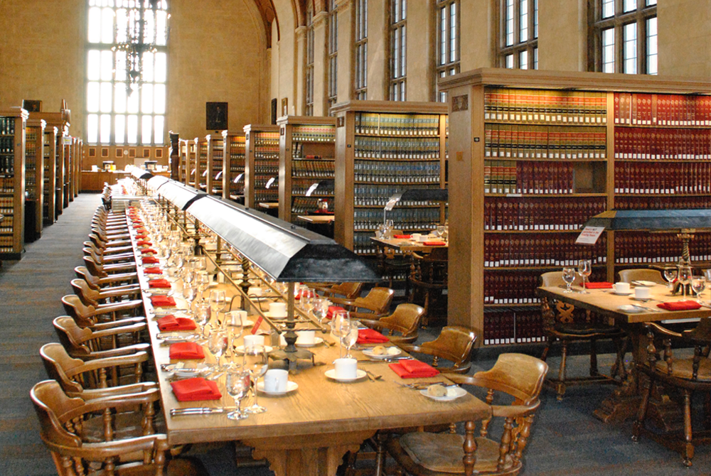 A photo of a long wooden table in the center of the Cornell Law Library that is set with red cloth napkins and bread plates.
