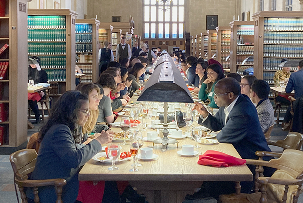 Photo of many people seated on either side of a long retangular table in the middle of the Cornell Law School library for a special feast.