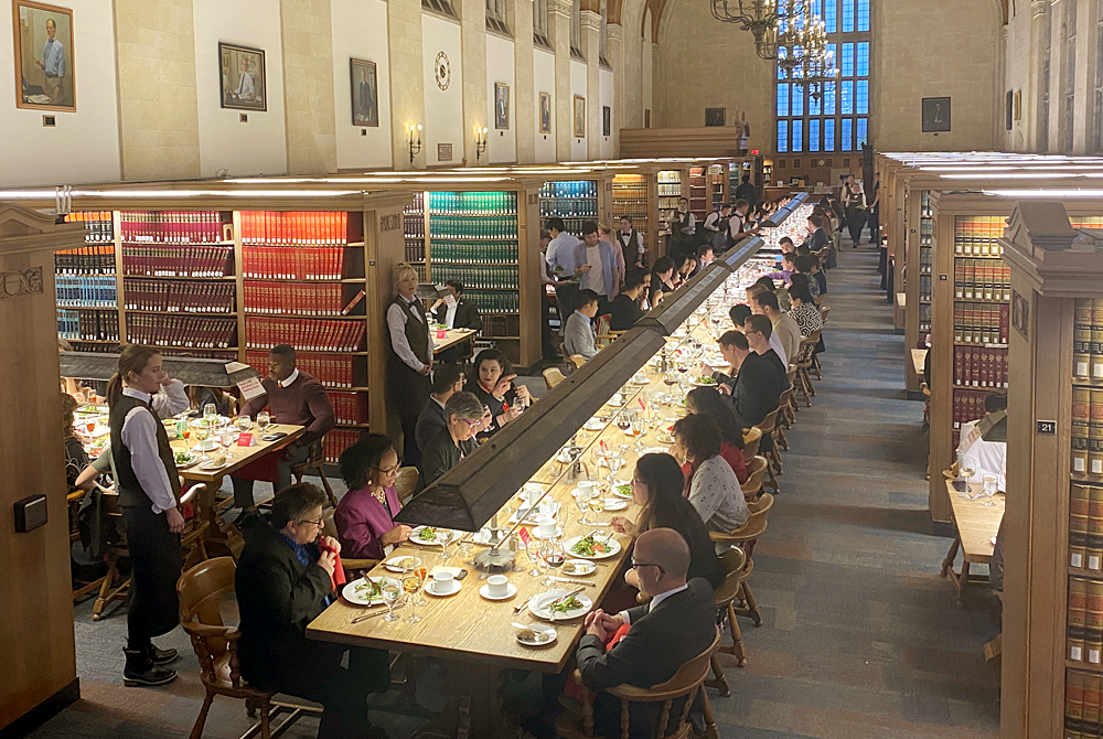 An arial photo showcasing the Cornell law Library with a feast laid out on the long tables down the center. Faculty, staff, and students are all seated and conversing and eating together.