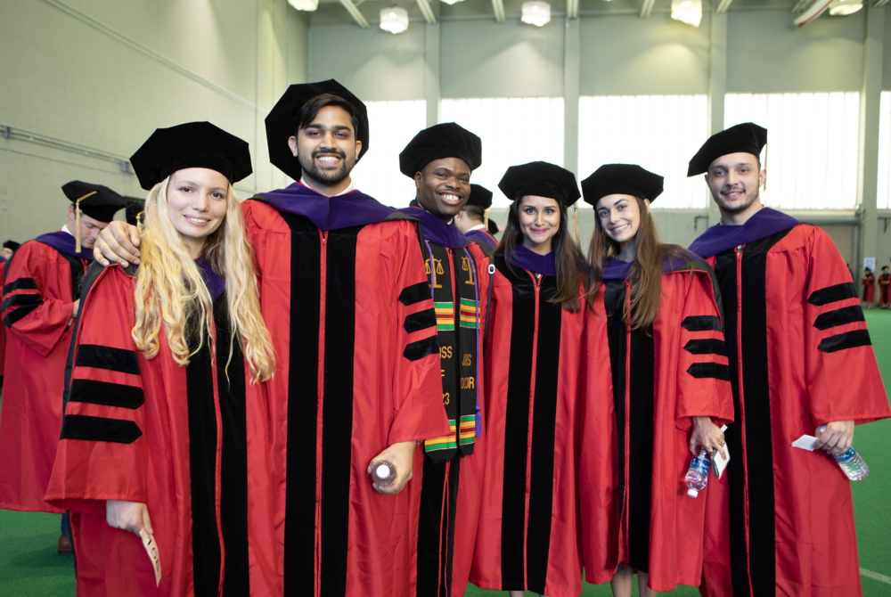 Several students wearing full graduation regalia are standing in a line and smiling at the camera. They have black velvet graduation caps on