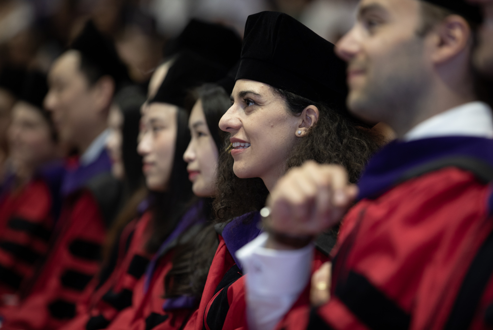 Several graduates wearing red and black regalia are sitting in the arena and looking up at a speaker. Everyone is blurred except for a woman who is smiling and has brown hair.