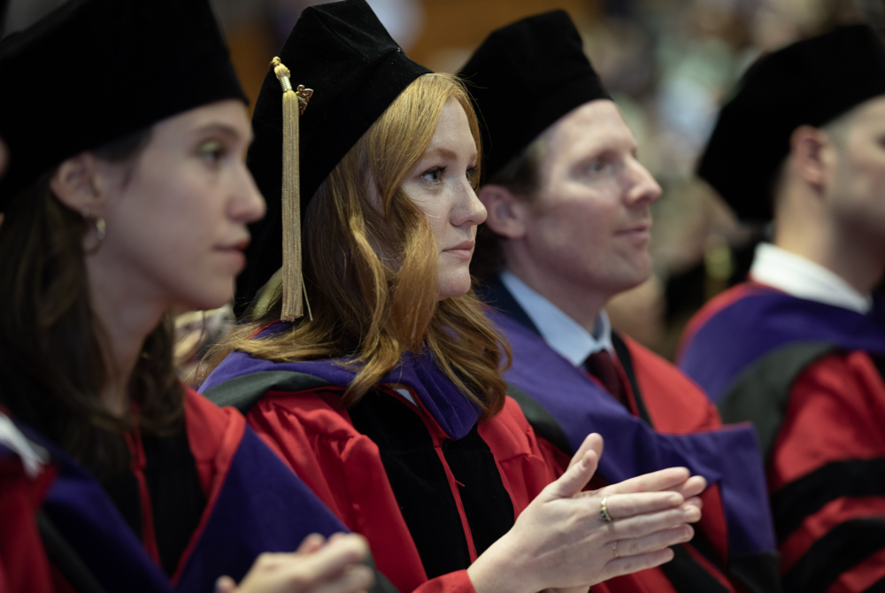 Students are wearing graduation regalia and clapping while looking up at the stage