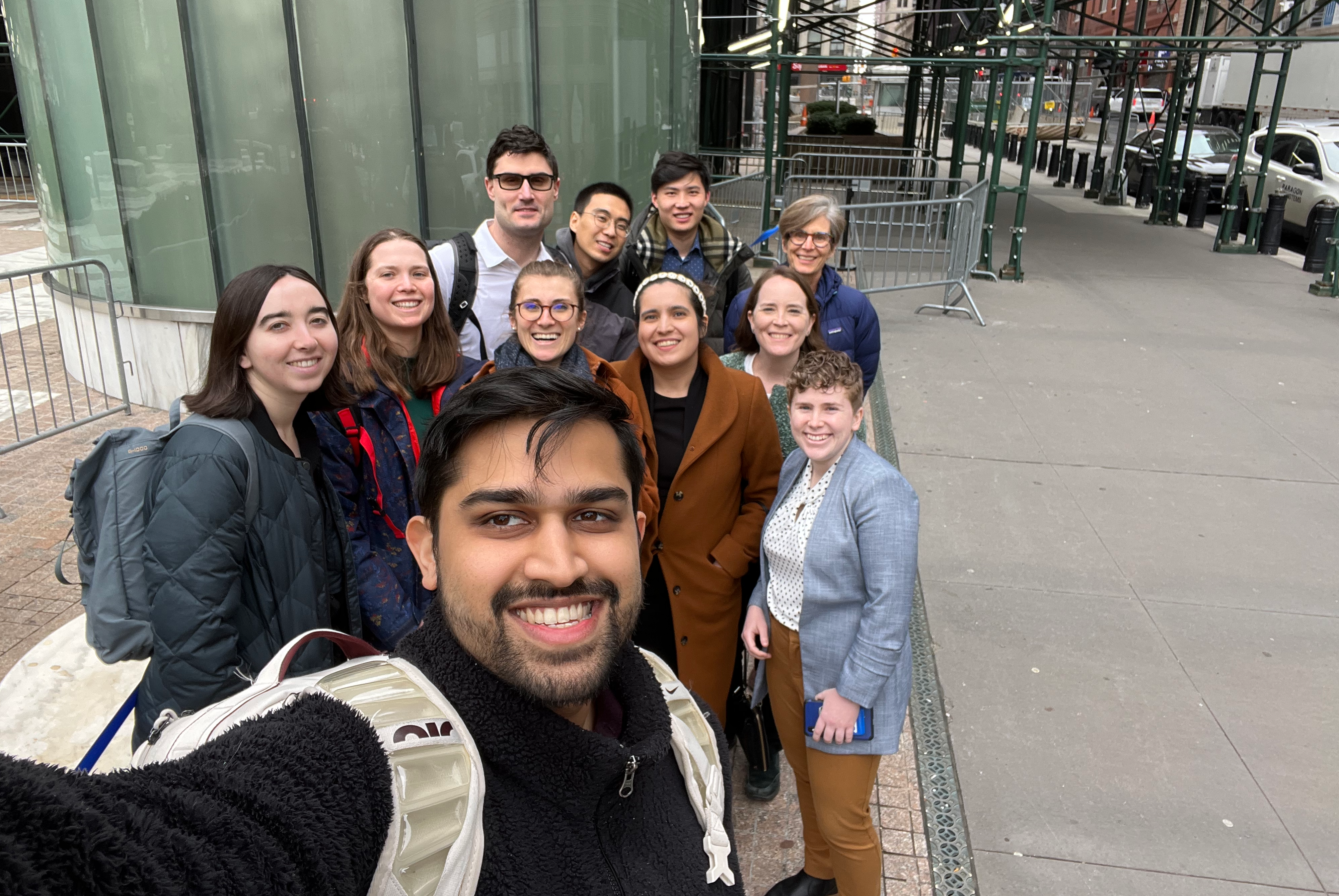a selfie photo of a group of Cornell Law students who are outside in a group on the streets of New York City. They are all looking up at the camera and smiling. The side of a green building is behind them.