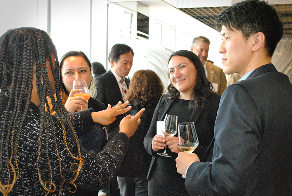 four students, all wearing black business attire are standing in a group during a cocktail reception. They are engaged in conversation and holding wine glasses in their hands.