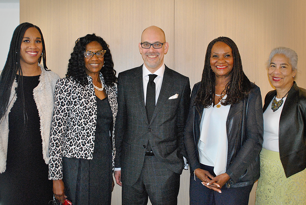 five people standing in a row facing the camera for a posed photo. On the left side are two women, a man in the middle wearing a suit, and two women standing on the right side of him as well.