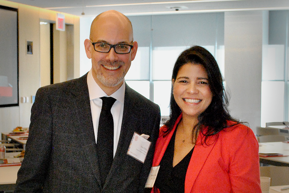 Image of two people smiling at the camera during an event in a large conference room. The gentleman on the left is bald, wearing glasses and is dressed in a suit and tie. The woman on the right has shoulder length brown hair and is wearing a black dress with a bright red blazer over top.