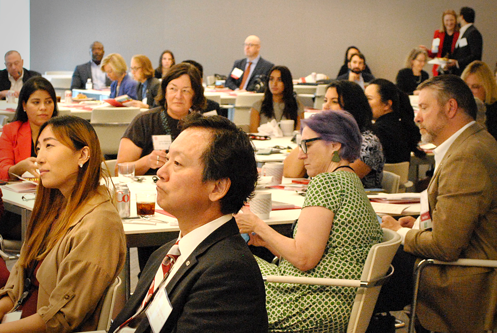 A crowd shot of several people sitting at large round tables and facing to the left. They are attending a conference and wearing name tags. The crowd is made up of various ethnicities, races, and genders.