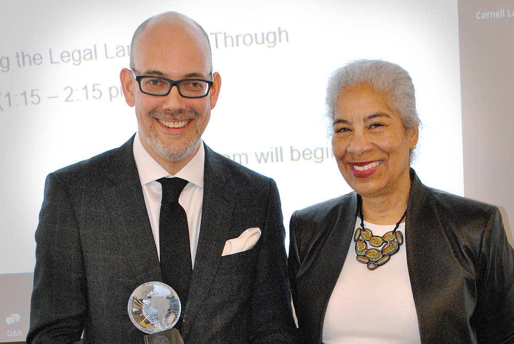 Two people face the camera smiling. On the left is a man wearing a black suit and tie, he is bald, wearing glasses and holding a glass trophy award. On the right is a woman with white hair, a chunky necklace and a white shirt with black leather blazer overtop.