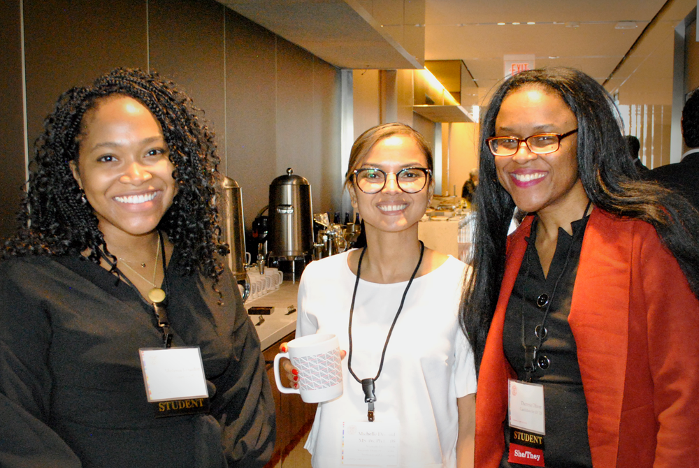 Three women are standing in a hallway, wearing nametags on lanyards and smiling at the camera. All three women are dressed in business casual attire. The woman on the left is wearing a black dress, the woman in the middle is wearing a white blouse and glasses, and the woman on the right is wearing glasses, a black dress, and a bright red blazer.