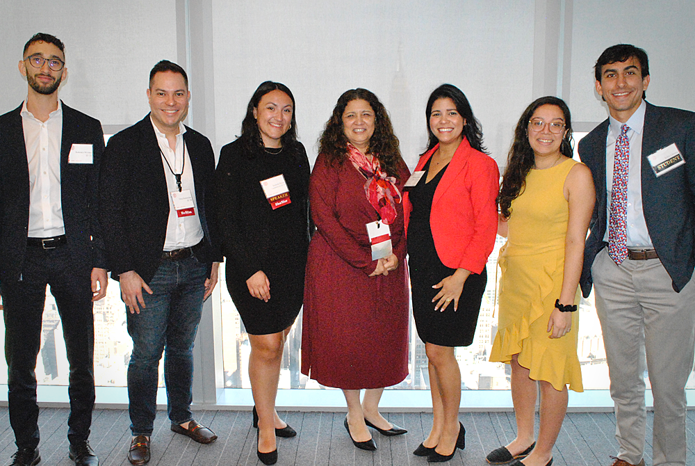 Seven people are standing in a line, facing the camera. They are all wearing business attire with nametags on lanyards around their necks. A large window with blinds drawn is behind them.