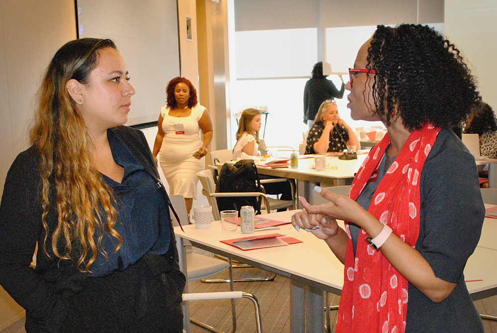 Two women are facing each other in a candid, action shot during a conference in a business suite. The women are engaged in conversation and there are people in the distance mingling in the room with large, round tables.