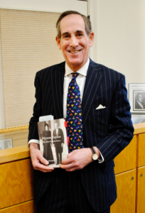 Photo of a man wearing a black suit and tie holding a copy of a book with a black and white cover. He is smiling at the camera and standing in front of a wooden display case.