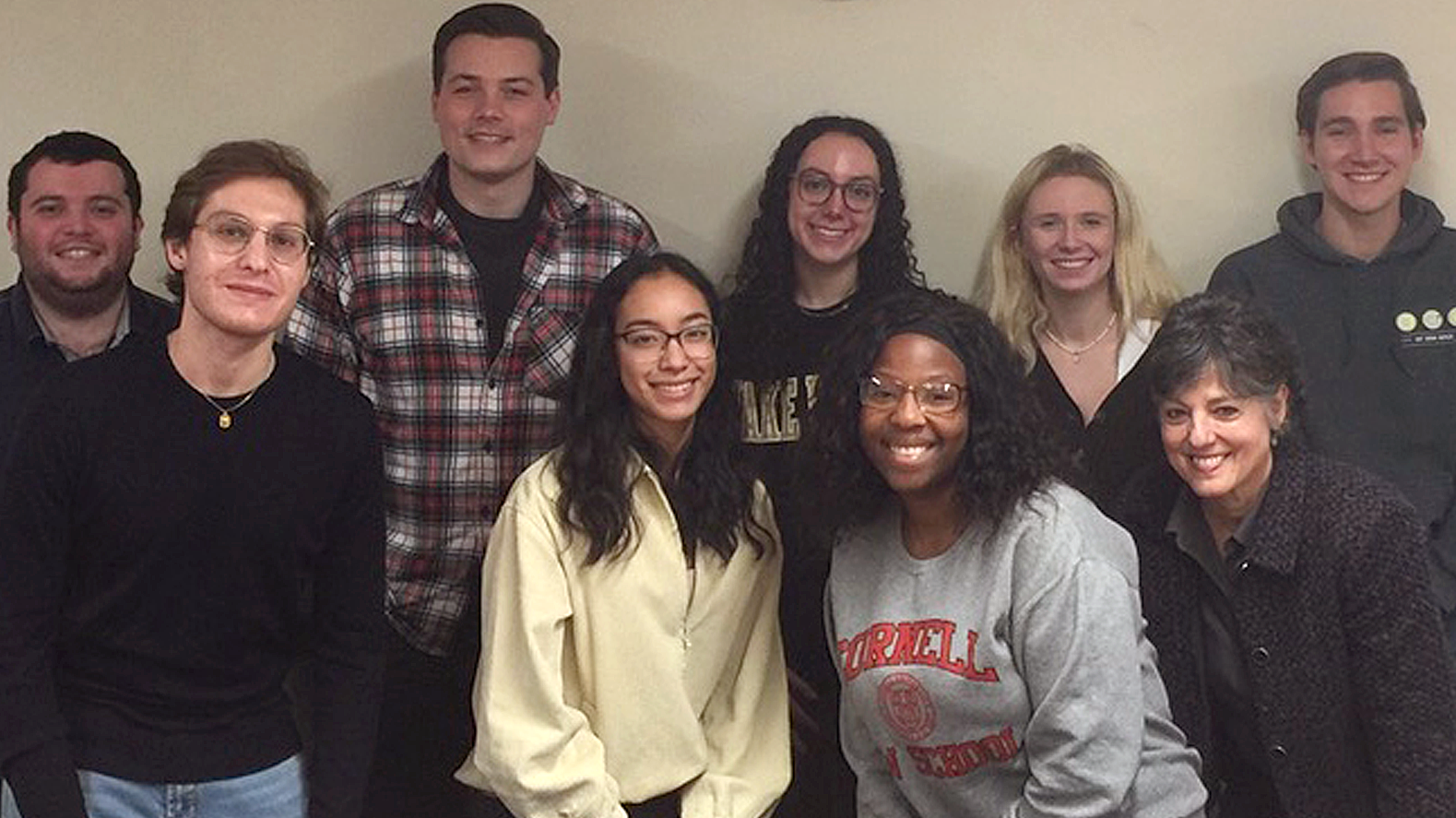 Image of 9 students from the labor law clinic after winning a settlement. They are all huddled together looking at the camera and smiling. A tan wall is behind them