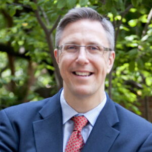 Headshot of Frank Pasquale wearing a suit and clear framed glasses. There are green tree branches behind him.