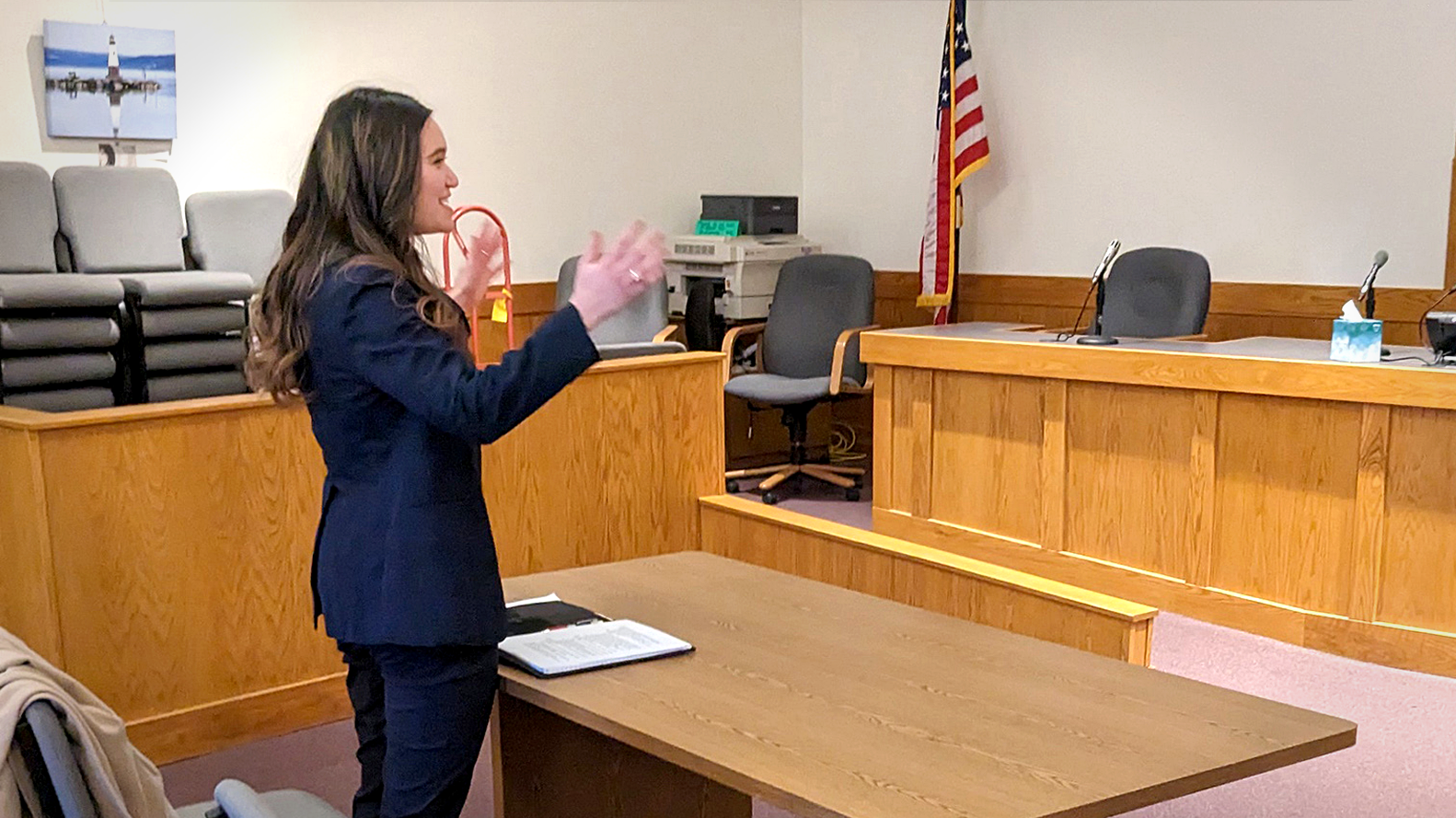 Photo of a woman wearing a navy suit standing in a court room behind a wooden table. She is gesturing her hands in the air towards the right side of the frame and appears to be speaking.