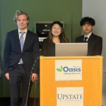 Photo of 5 people standing in suits behind a podium. The podium has two signs on the front that are white and have logos on them for Oasis and Upstate