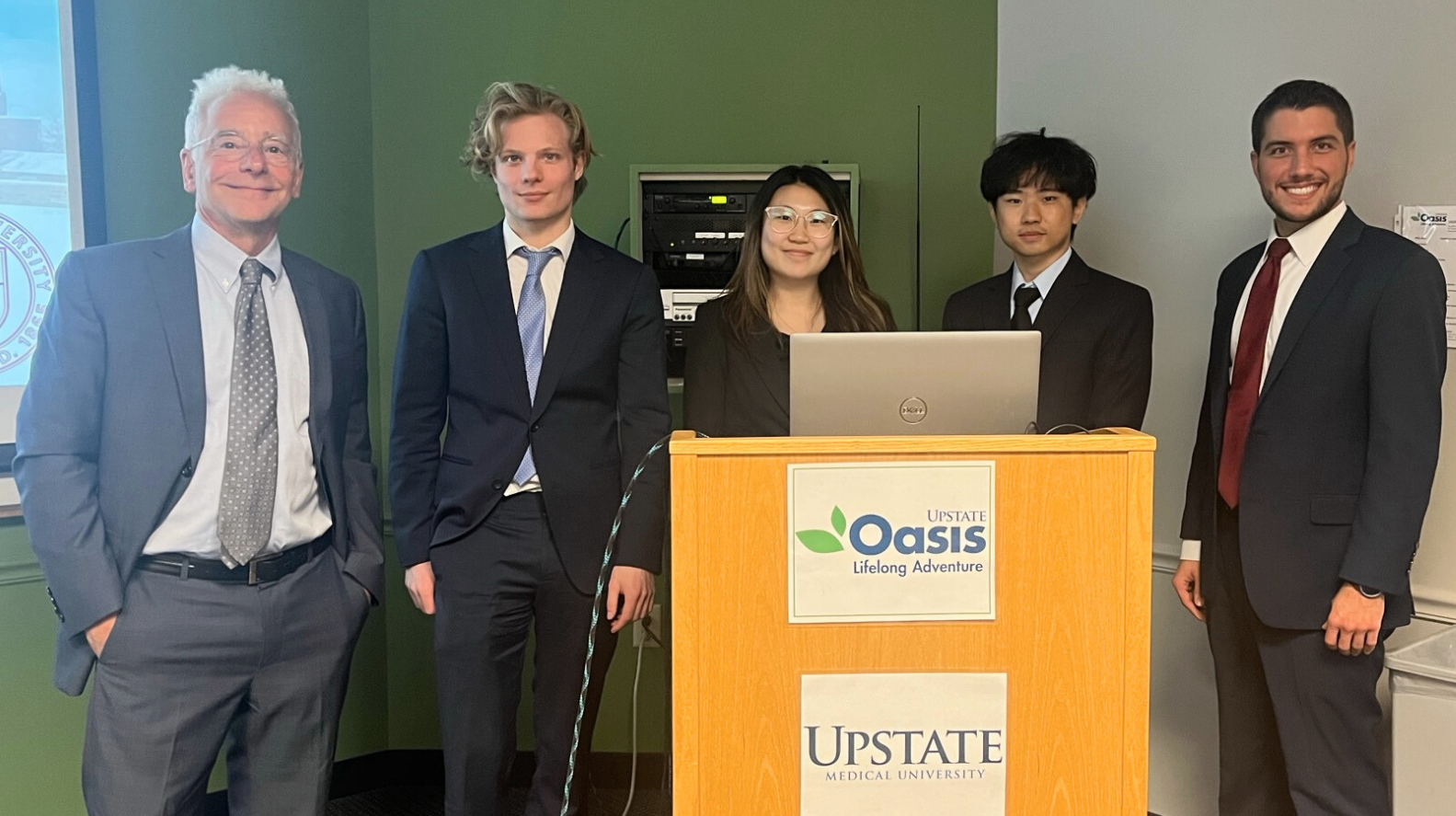 Photo of 5 people standing in suits behind a podium. The podium has two signs on the front that are white and have logos on them for Oasis and Upstate