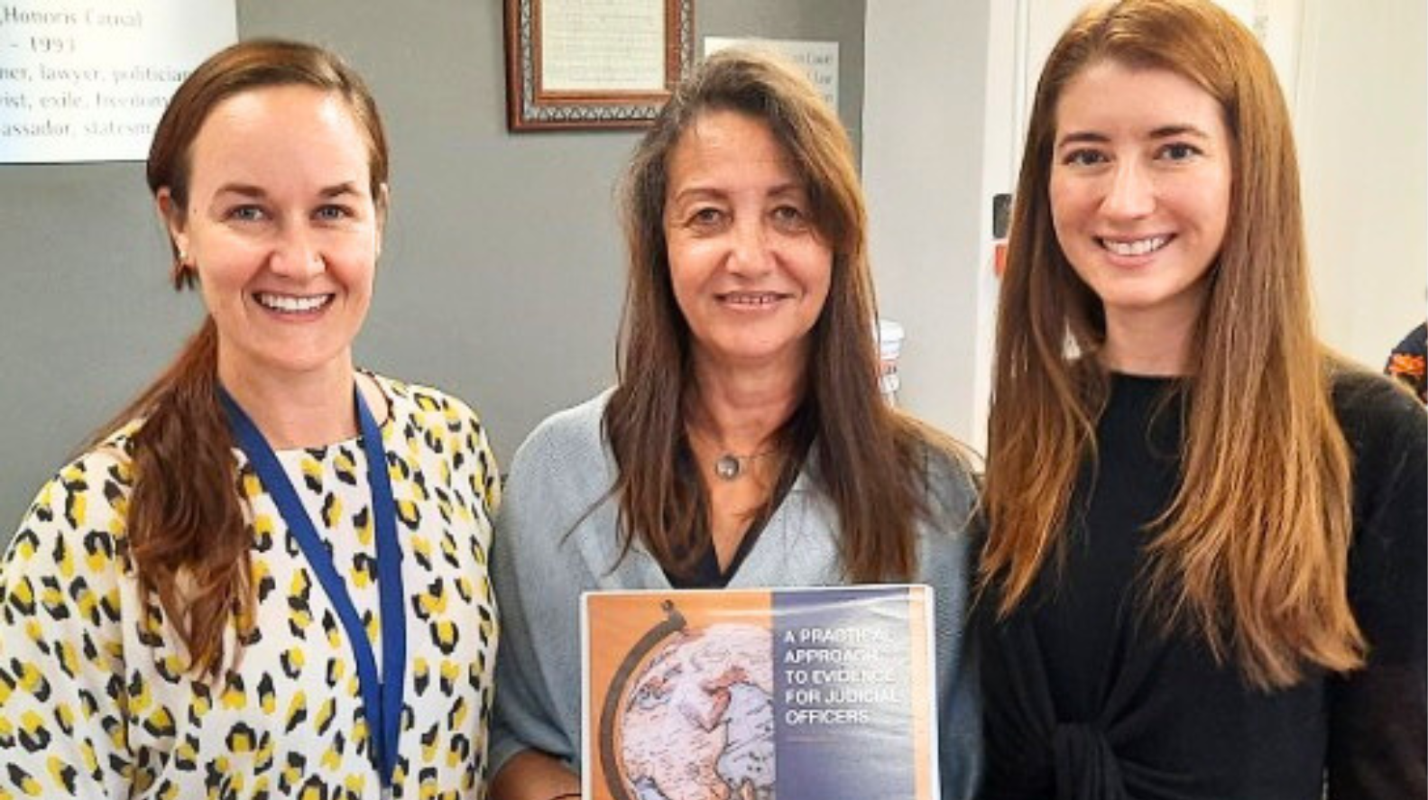 Three women with long hair are standing in a row and smiling at the camera. The are holding a sign in a plastic sleeve that has a photo of a globe on it. There is a white and pale blue wall behind them.