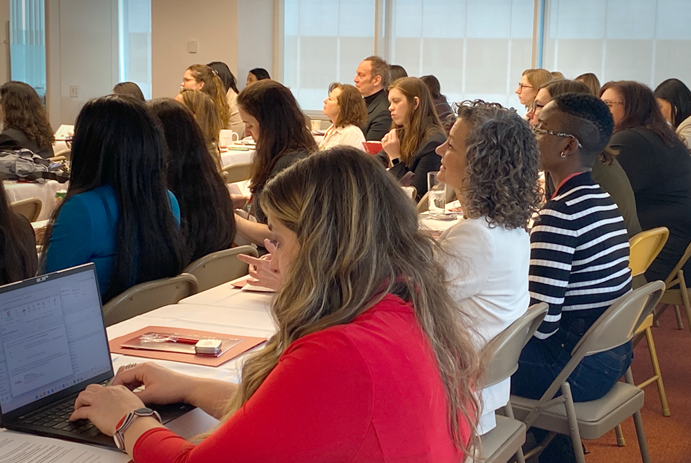 Several women sitting at long tables in a conference room are facing away from the camera and looking to a speaker off camera.