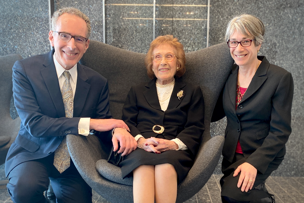 three people are facing the camera and smiling. On the left is a man kneeling and resting his hand on the arm of an elderly woman who is seating in the middle in a grey arm chair. On the right is a woman also kneeling and embracing the chair for a posed photo.