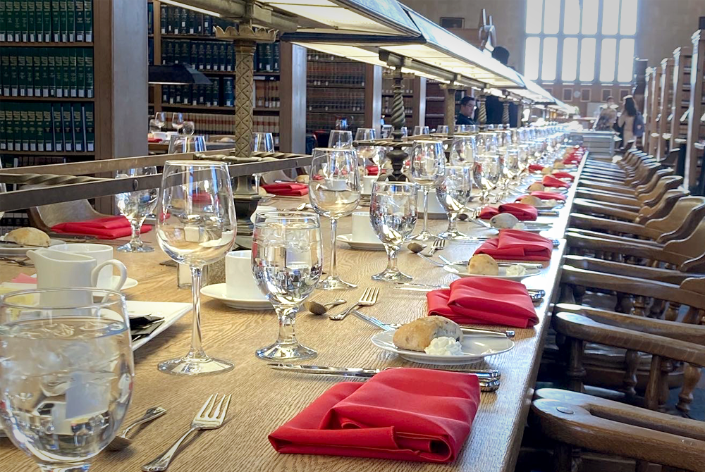 Photo of place settings down a long table. Red cloth napkins are folded at each place setting and empty water and wine glasses are paired in front. along the table in the center are large lamps for studying and library stacks are blurred in the background.