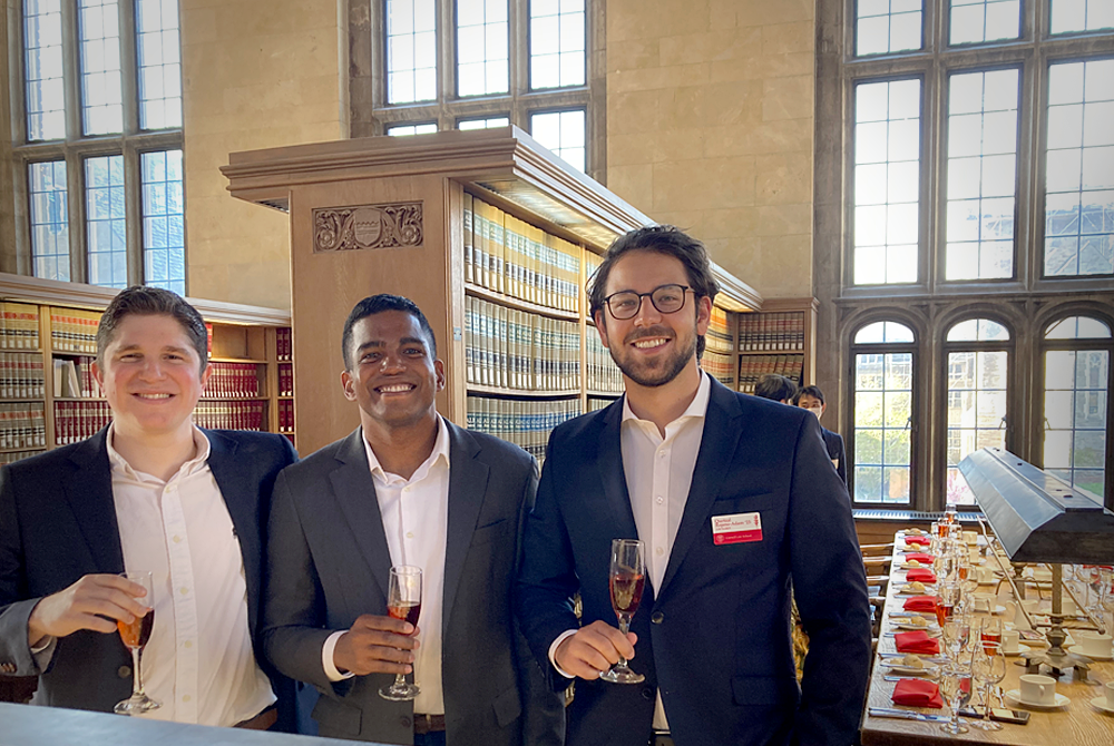 Three men standing in suits in the library at Cornell Law School. They are each holding champagne glasses in their right hands