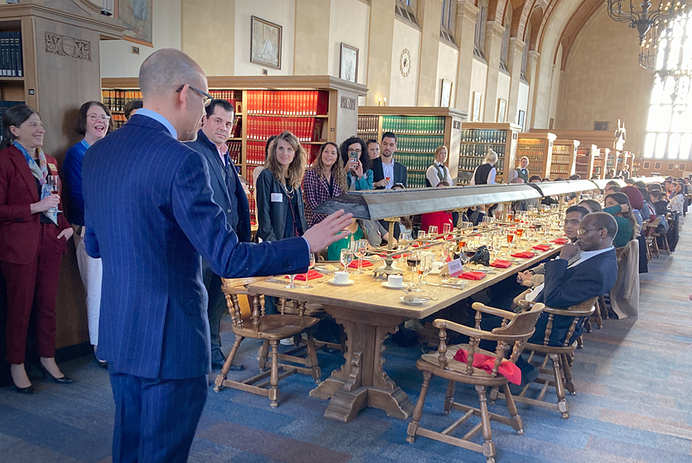 A photo of a gentleman wearing a blue suit who is standing in front of a long table in the law library. His hands are raised as he is giving a speech and welcoming remarks to many people seated and standing down the center isle.