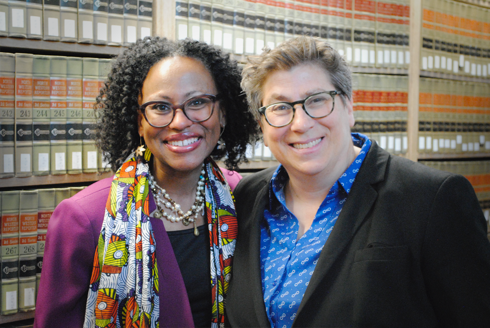 Two women who are wearing business attire and standing in a library stack in front of books.