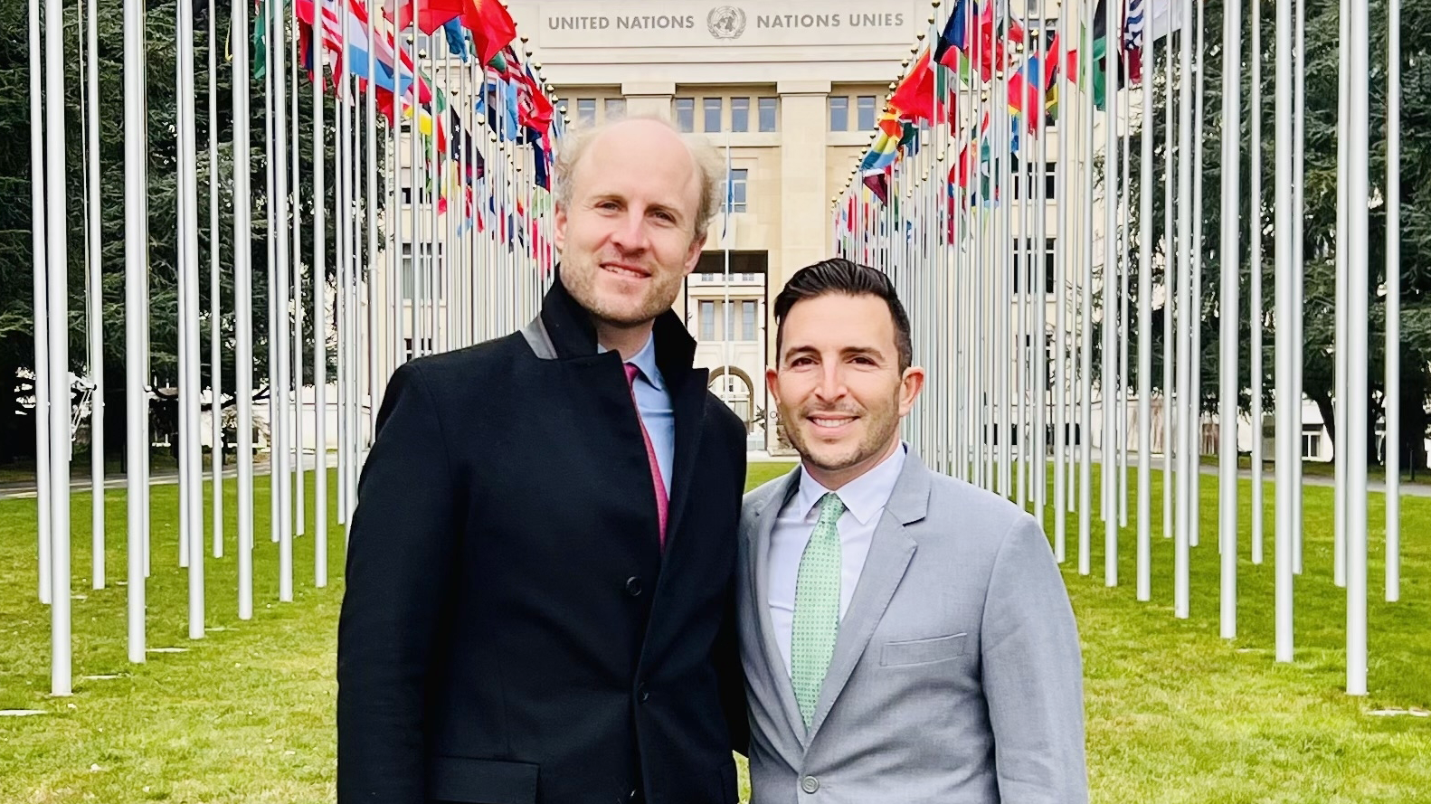 two people wearing suits are standing alongside each other. Bright green grass is on the ground and long rows of international flags line the yard behind them. They are standing at the UN