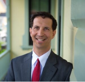 Headshot of Dan Berger. He is wearing a charcoal grey suit with a bright red tie. Behind him is a balcony with white railings