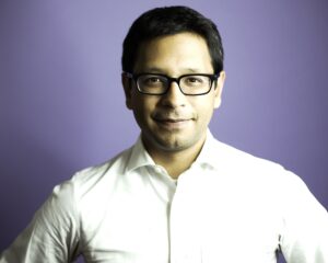 Headshot of a man wearing a white suit shirt and black frame glasses. He had dark hair and is standing in front of a purple background