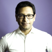 Headshot of a man wearing a white suit shirt and black frame glasses. He had dark hair and is standing in front of a purple background