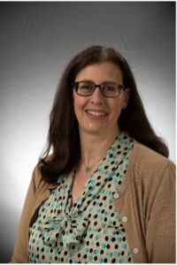 Photo of a woman wearing a beige cardigan and a teal blouse with a grew background behind her. She has long brown hair and glasses.