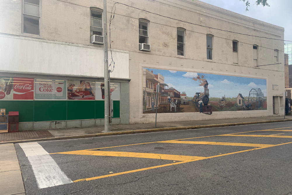 A photo of the side of a white brick building in Alexandria Louisiana with a large mural painted on the side that depicts a train.