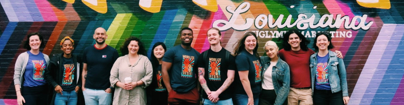 Photo of large group of people standing in front of a colorful mural painted on a wall behind them in a city in Louisiana.