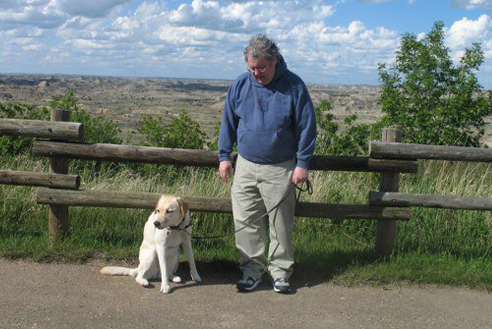 A man wearing a blue sweater is looking down at a yellow lab dog. They are outside with blue skies and green grass behind them.