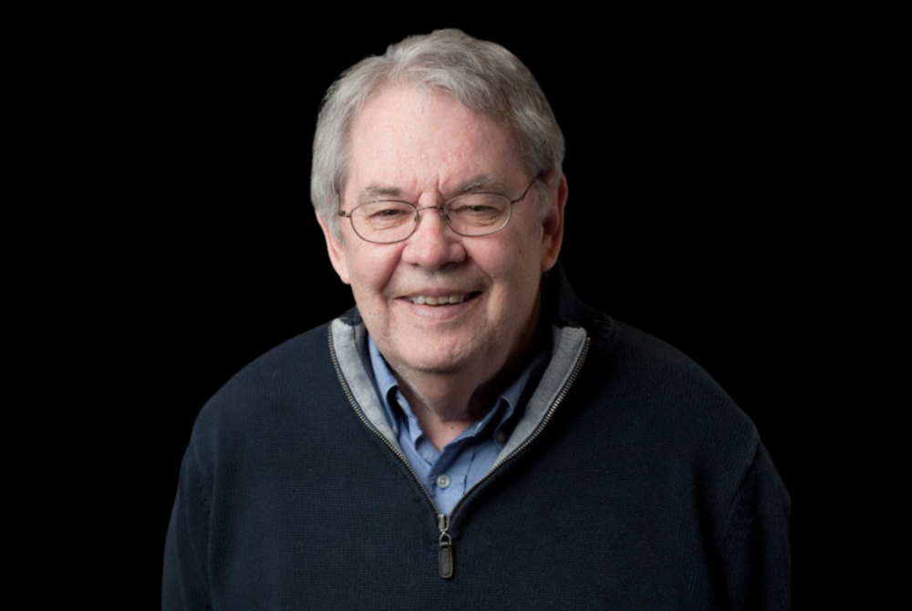 Portrait of Steve Shiffrin wearing a blue button down shirt and a navy sweater over top. He is smiling and wearing glasses. A black background is behind him.