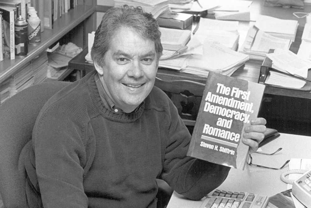 Photo of a man holding a book in his right hand. The title reads The First Amendment Democracy and Romance. He is sitting at a desk wearing a sweater. he is smiling at the camera