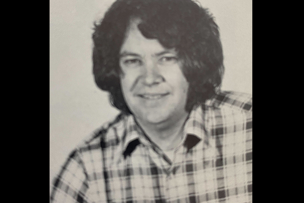 Black and white portrait of a man wearing a plaid shirt. He has full, shaggy hair and is smiling at the camera